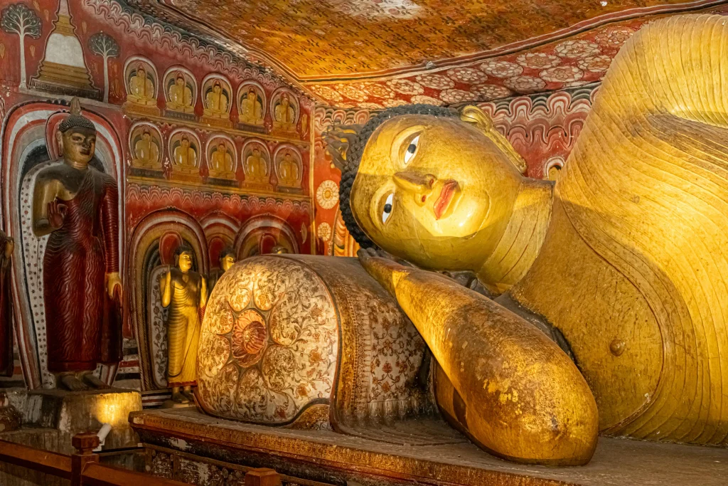 Golden reclining Buddha statue inside the ancient cave shrine at Dambulla Cave Temple in Sri Lanka with painted Buddhist murals and statues.